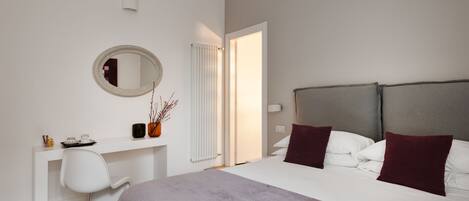 A modern bedroom with a large bed, a white desk, a chair, a mirror, and a radiator.