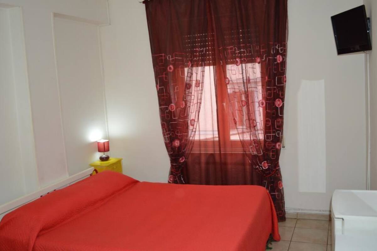 A bedroom with a bed, a bedside table with a lamp, a TV on the wall, and a window with a curtain.