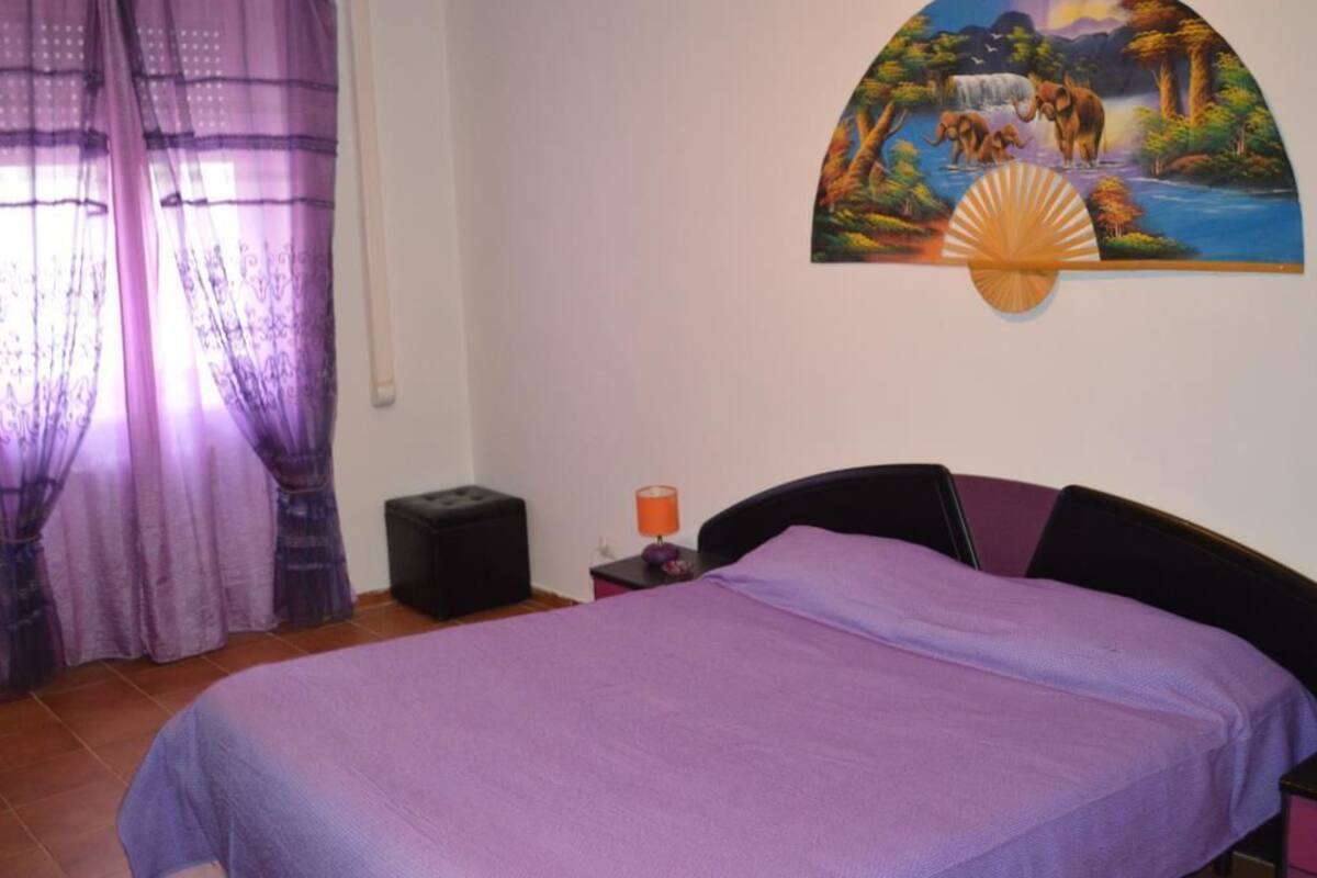 A bedroom with a bed, a nightstand, a lamp, a painting, and purple curtains.