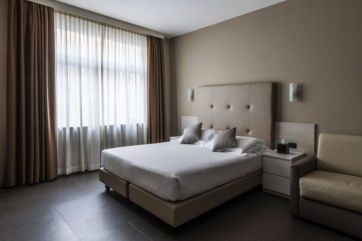 A modern hotel room with a large bed, a nightstand, a chair, and a window with sheer curtains.