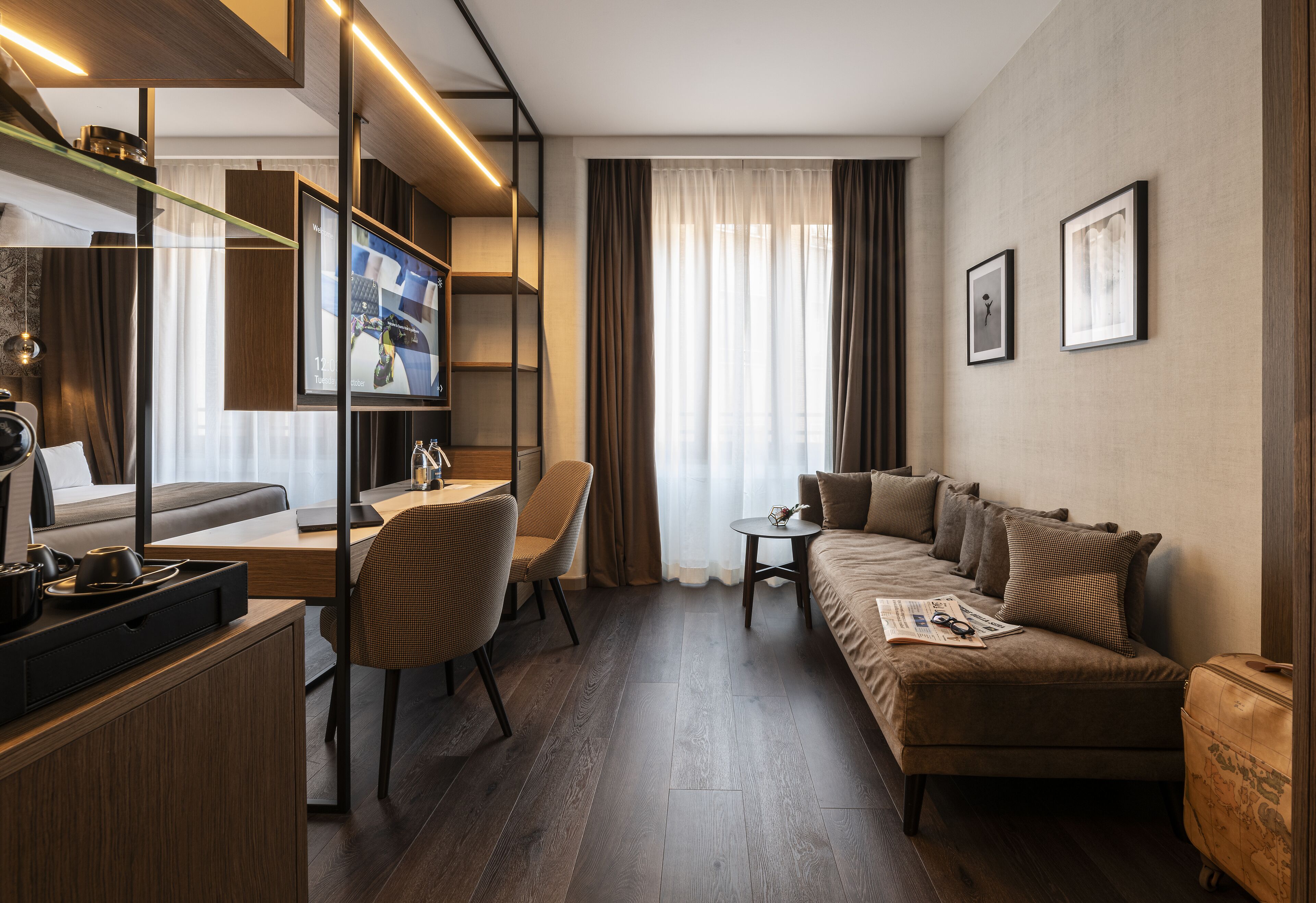 A modern hotel room with a wooden floor, a sofa, a desk with a chair, a small table, and a television.