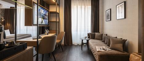 A modern hotel room with a wooden floor, a sofa, a desk with a chair, a small table, and a television.