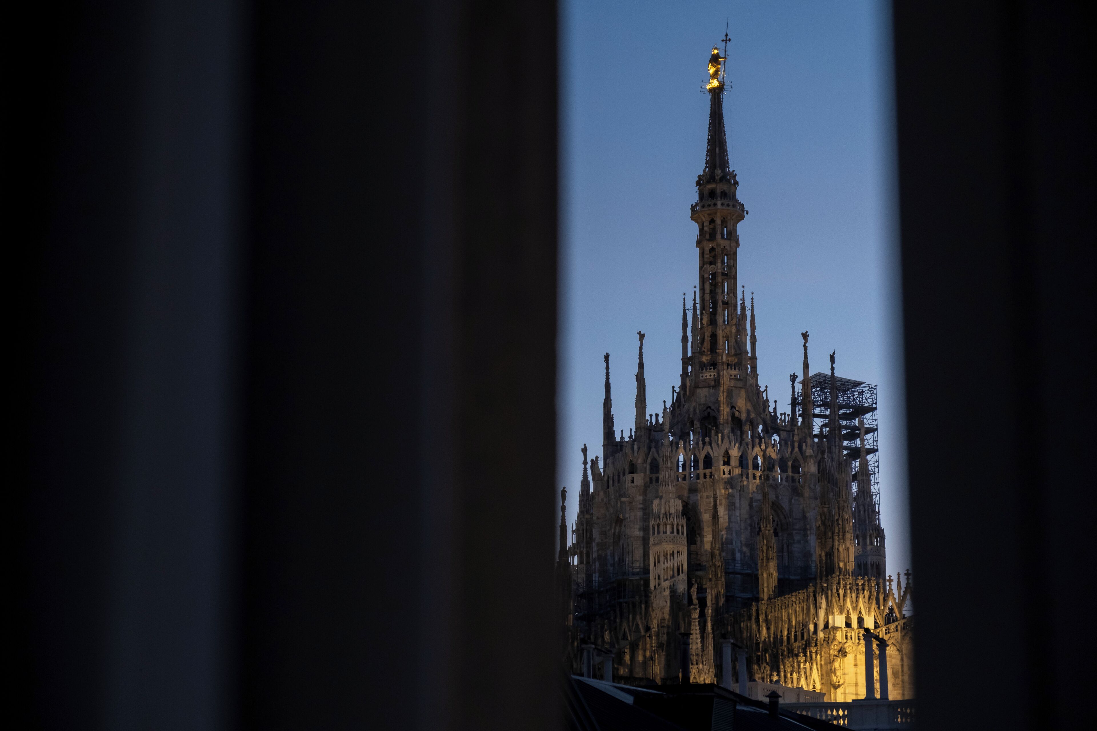 Una catedral gótica con intrincadas agujas y una estatua dorada en la cima, vista a través de una estrecha abertura.