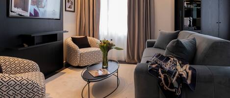 A modern living room with a grey sofa, patterned ottoman, and a small coffee table with a vase. A television is mounted on the wall, and there is a wardrobe in the background.