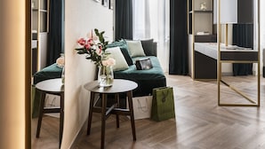Una habitación de hotel moderna con un sofá verde, una pequeña mesa redonda con un jarrón de flores, un espejo grande y un escritorio empotrado con estanterías.