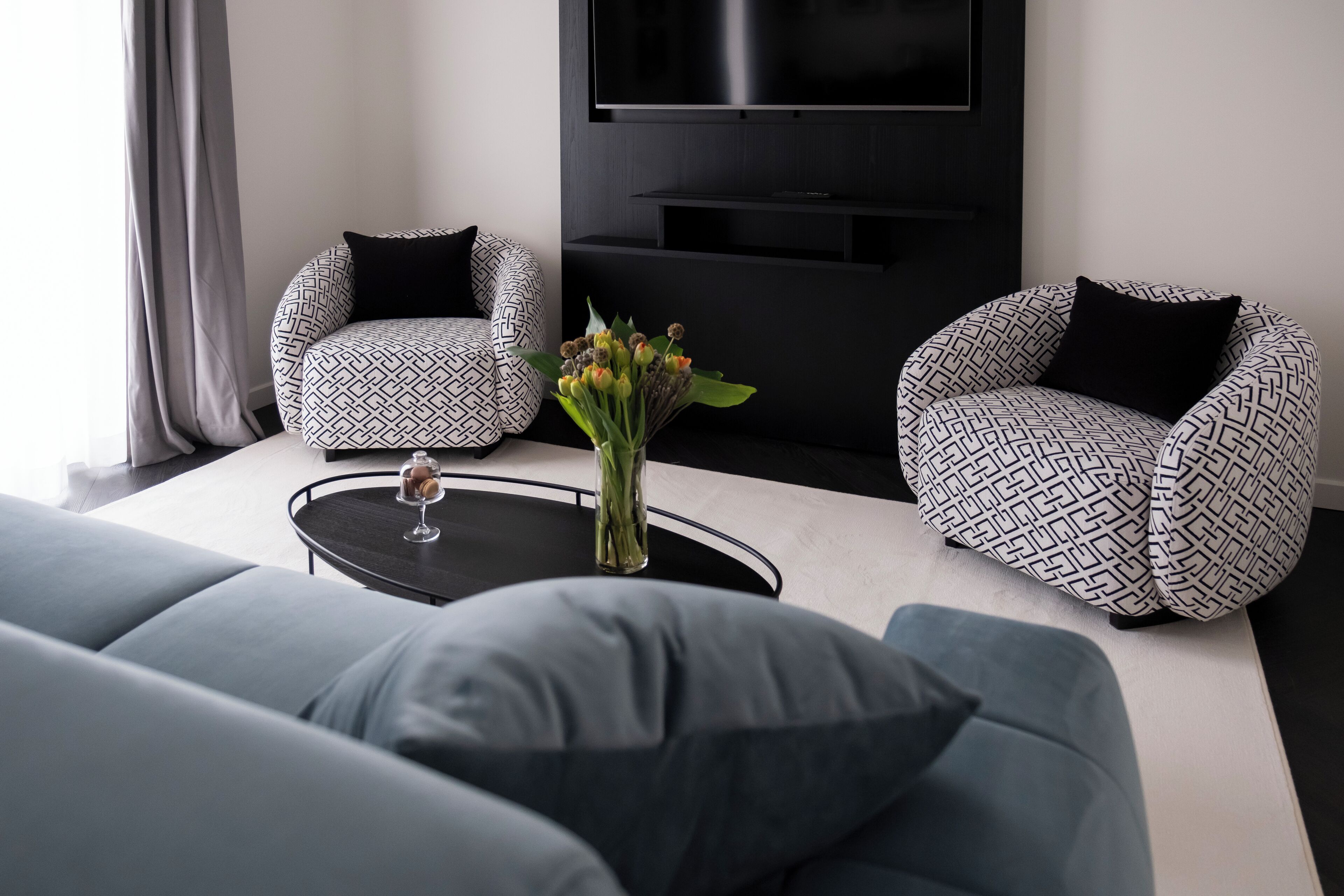 Un salon moderne comprenant un canapé, deux fauteuils à motifs, une table basse avec un vase de fleurs et un téléviseur à écran plat fixé au mur.