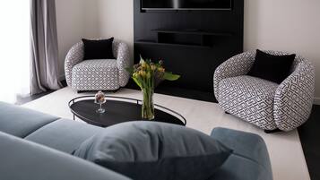 Un salon moderne comprenant un canapé, deux fauteuils à motifs, une table basse avec un vase de fleurs et un téléviseur à écran plat fixé au mur.