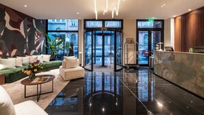 Un lobby moderno con un ascensor de vidrio circular, una recepción y una zona de estar con un sofá verde y una mesa de centro.