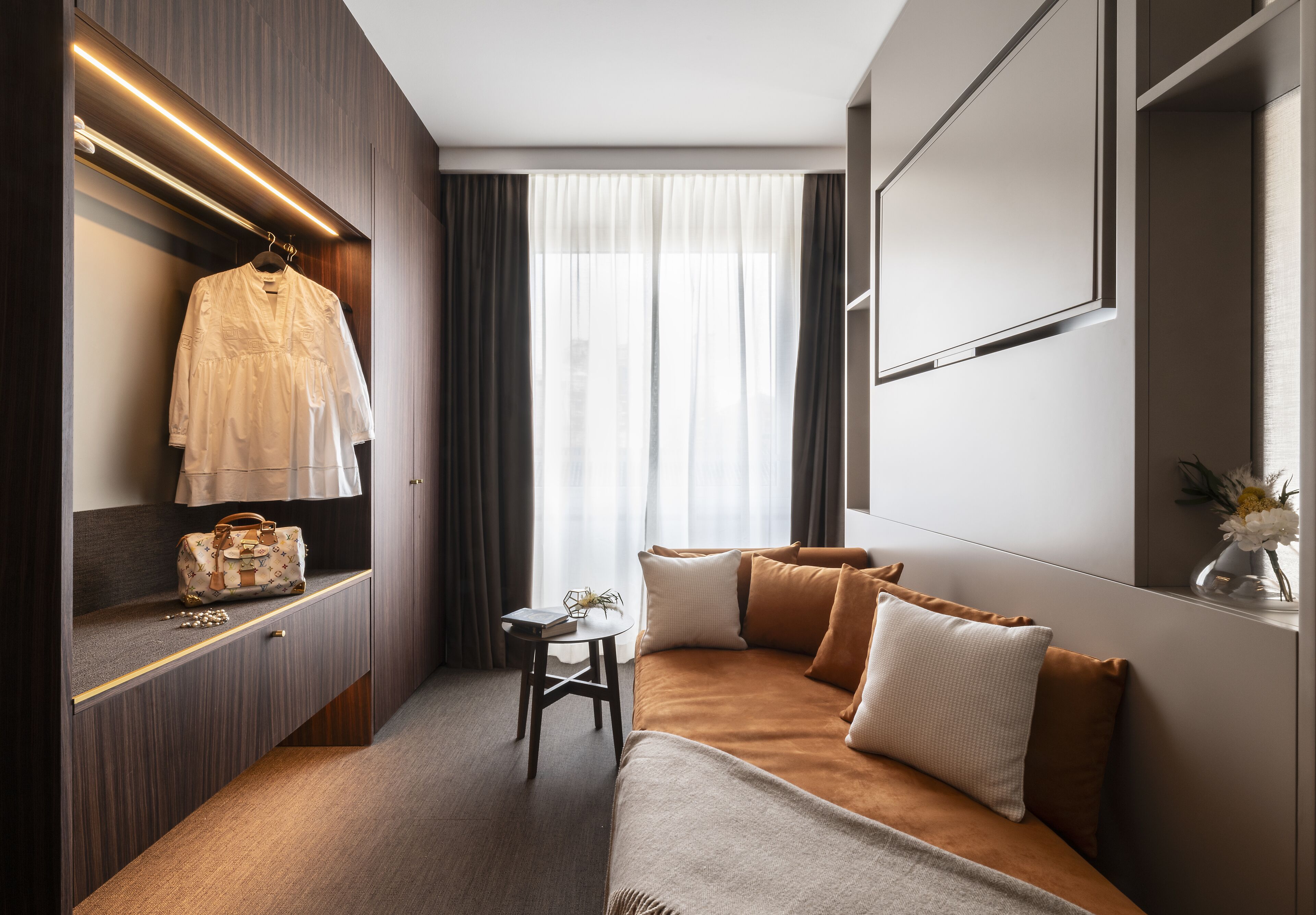 A modern hotel room with a bed, a vanity with a mirror, a hanging robe, and a small table with a vase.