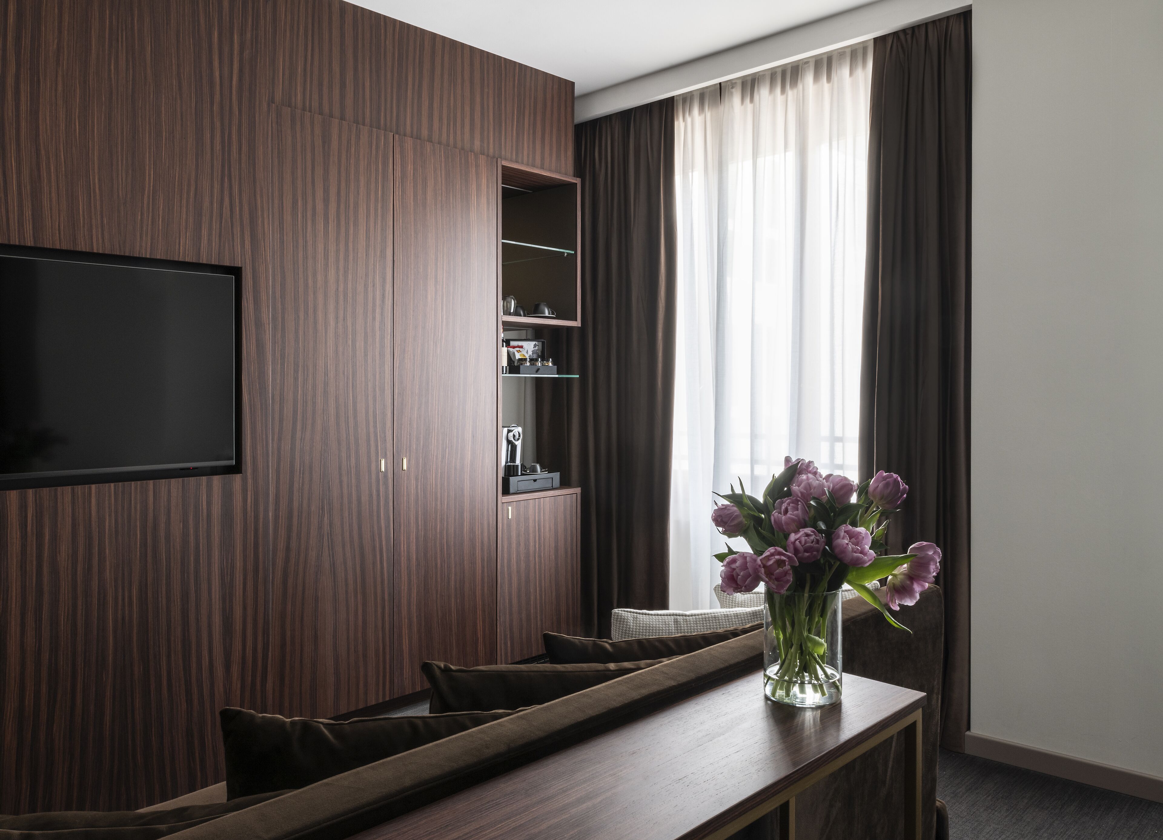 A modern living room with a dark wood panel wall, a flat-screen TV mounted on it, a glass coffee table with a vase of flowers, and a large window with curtains.