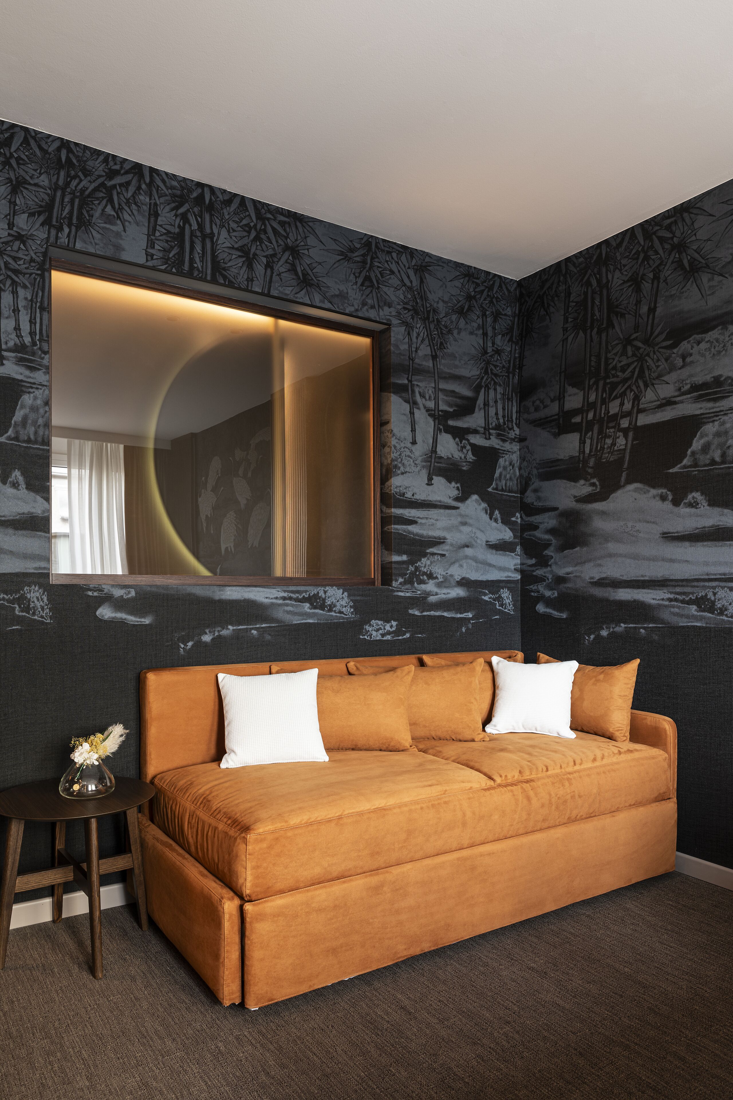 A room with a brown sofa, a round table with a vase, and a large mirror.