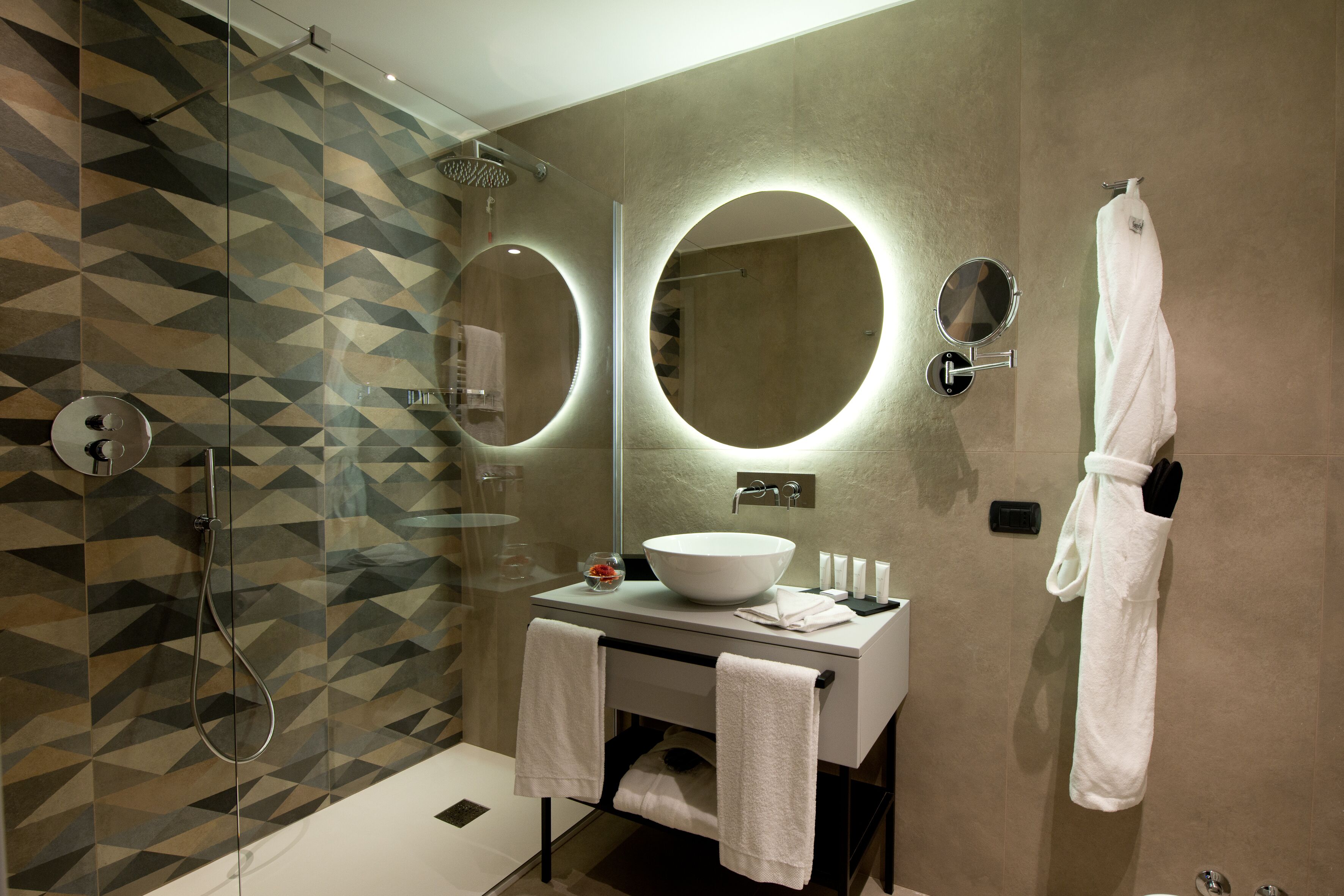 Une salle de bain moderne avec un mur recouvert de carreaux géométriques, un grand miroir rond, un lavabo blanc et une douche à parois de verre.