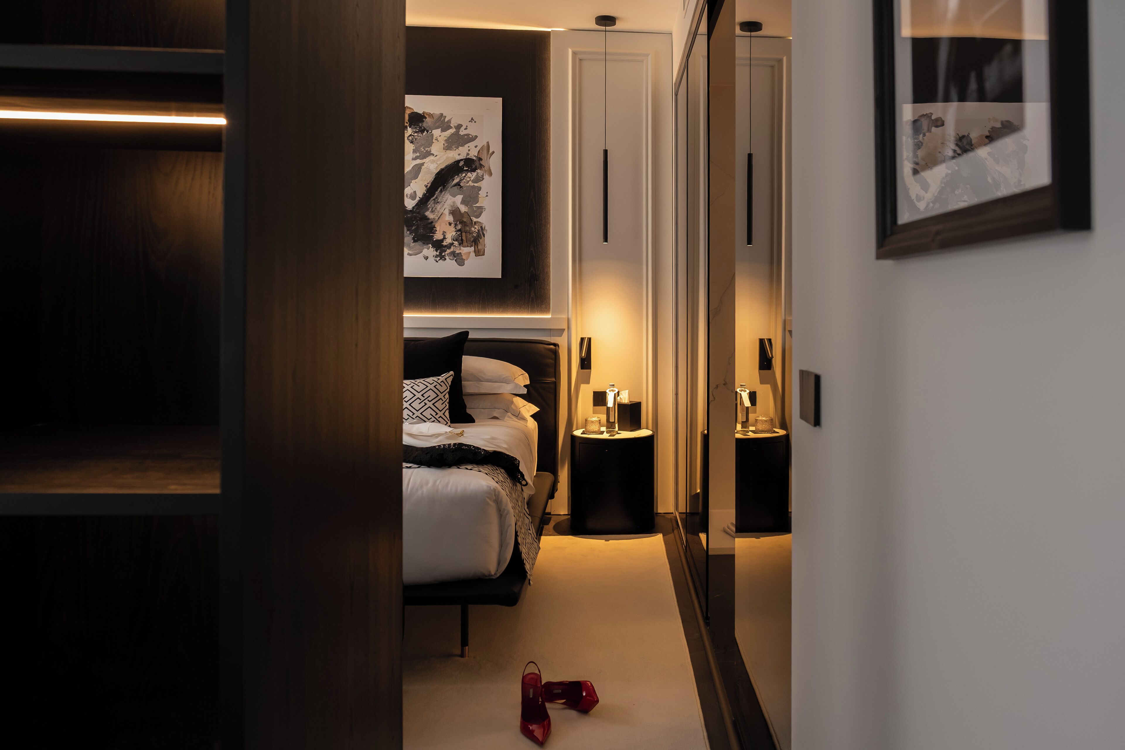 A modern hotel room with a neatly made bed, a bedside table, and a nightstand. There is a wardrobe with a mirror and a framed artwork on the wall.