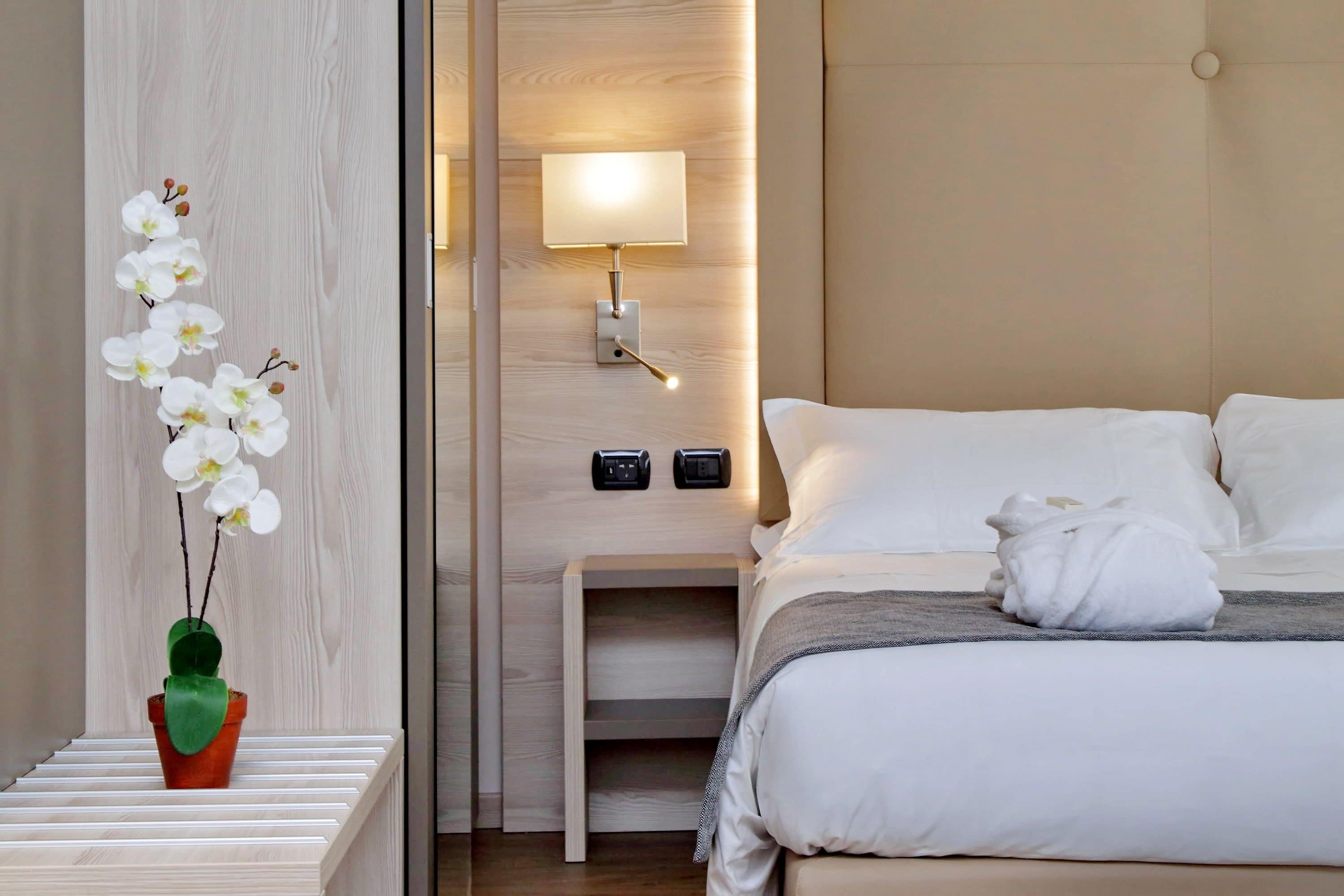 A neatly arranged hotel room with a bed, bedside table, lamp, and a potted orchid.