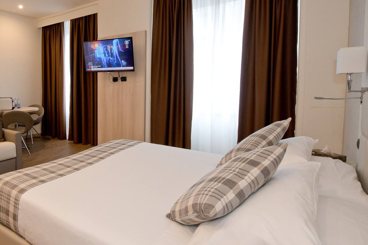 A hotel room with a bed, a sofa, a TV mounted on the wall, and a window with curtains.