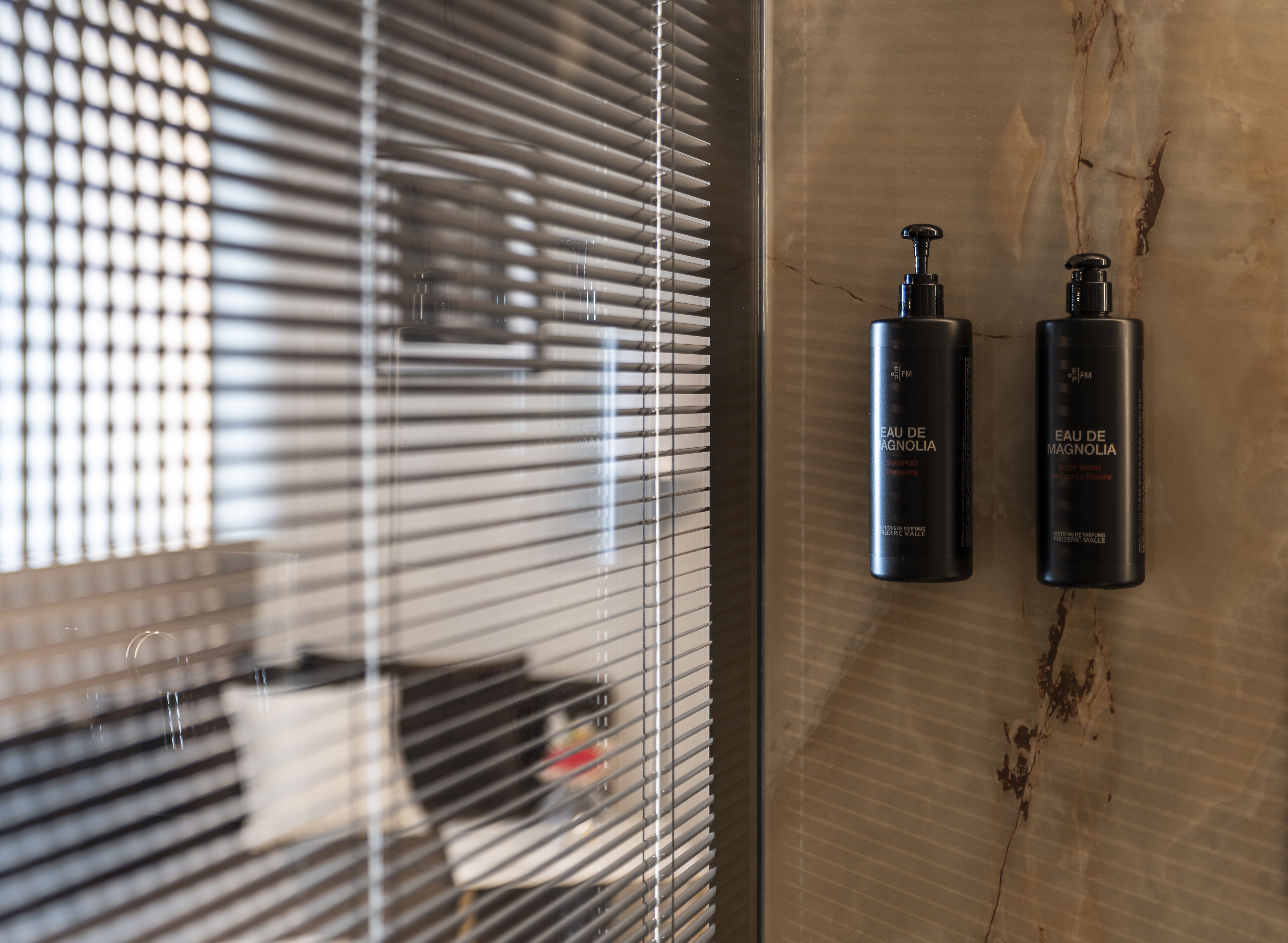 A bathroom with a marble wall, two black bottles of shampoo and body wash labeled 'EAU DE MAGNOLIA', and a window with blinds partially open.