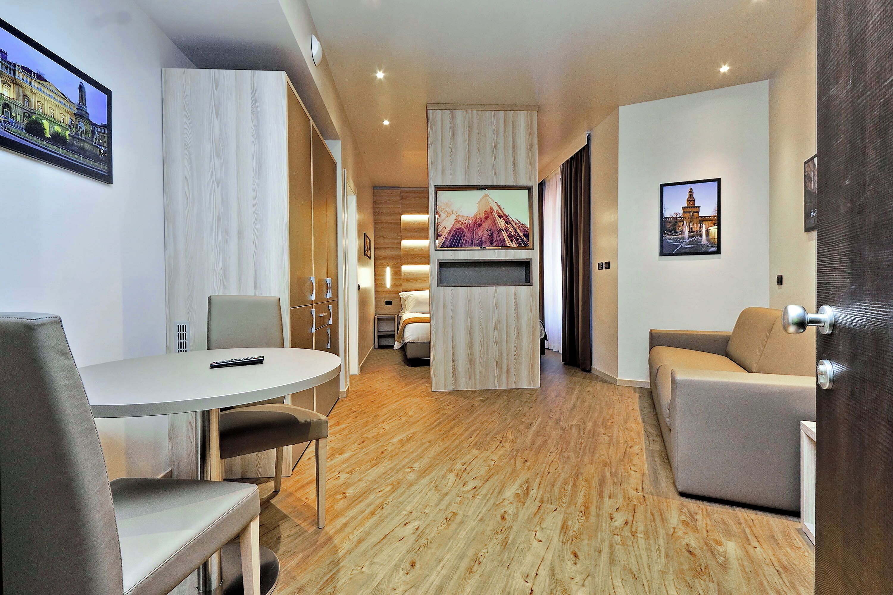 A modern hotel room with a wooden floor, a round dining table with chairs, a sofa, and a TV mounted on the wall.