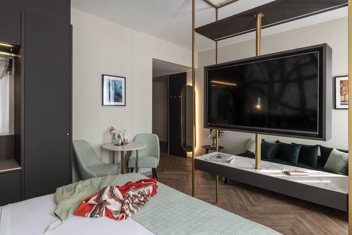A modern hotel room with a large bed, a TV mounted on a wall, a seating area with a sofa and chairs, and a small table with a vase.