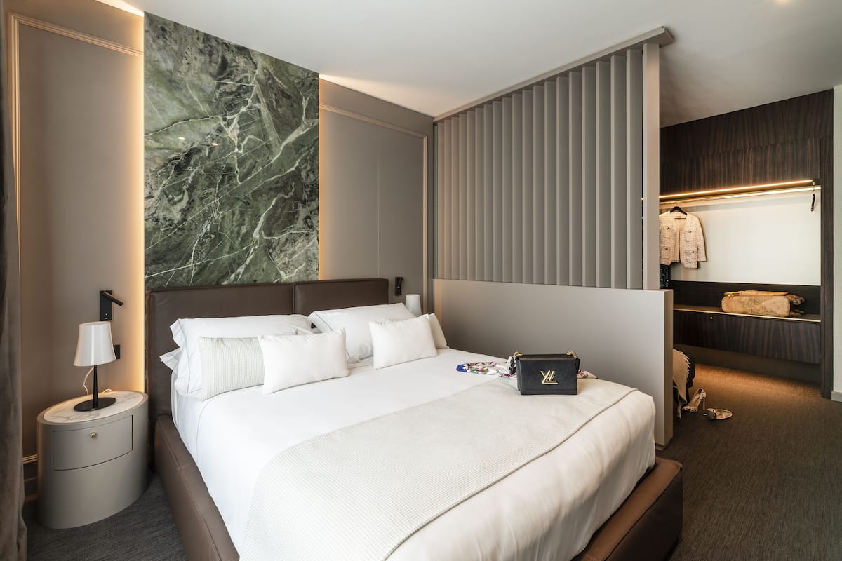 A modern hotel room with a large bed, a nightstand with a lamp, a marble wall, a built-in closet with a mirror, and a shoe rack.