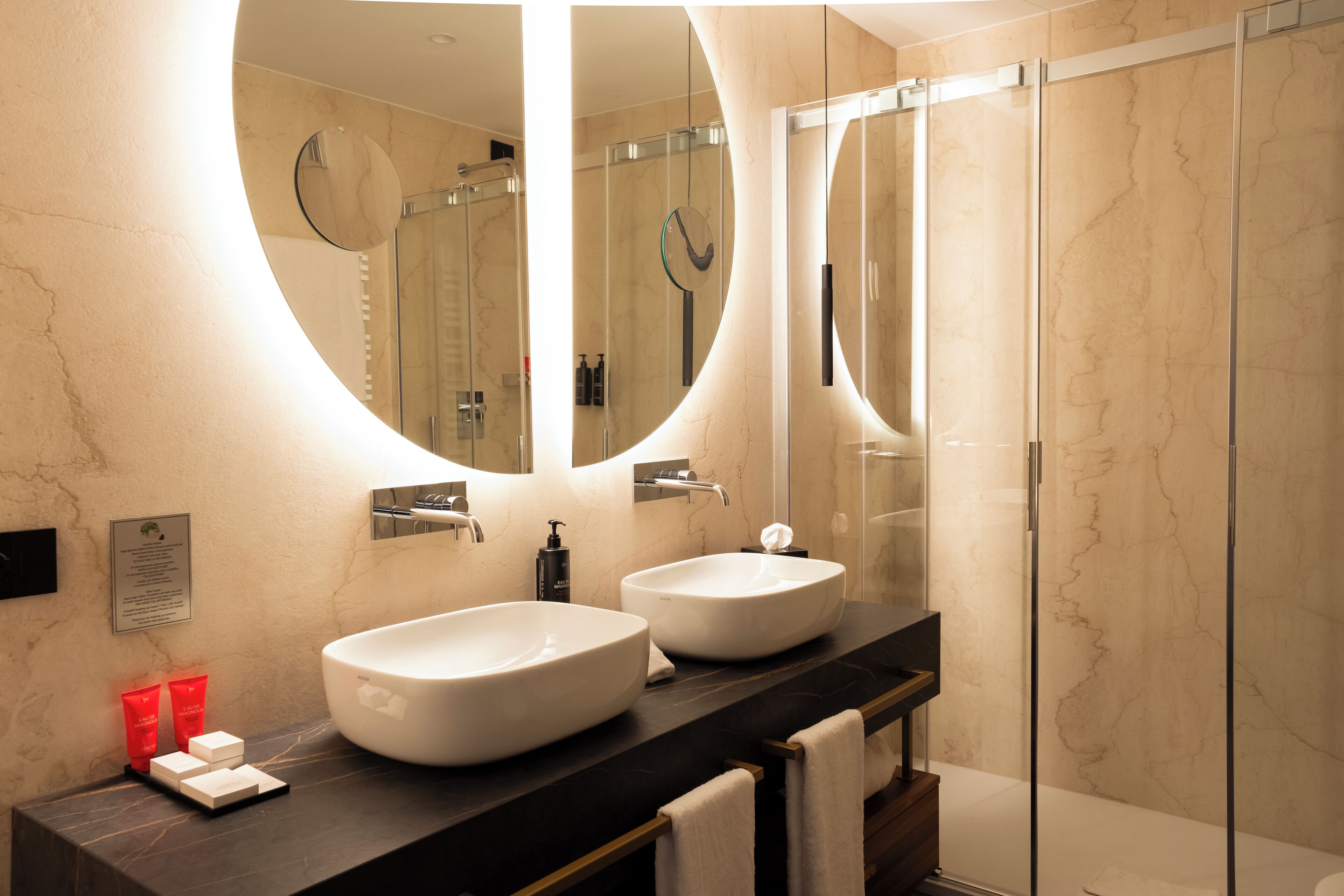 A modern bathroom with two large white sinks, a dark marble countertop, and a glass shower enclosure.