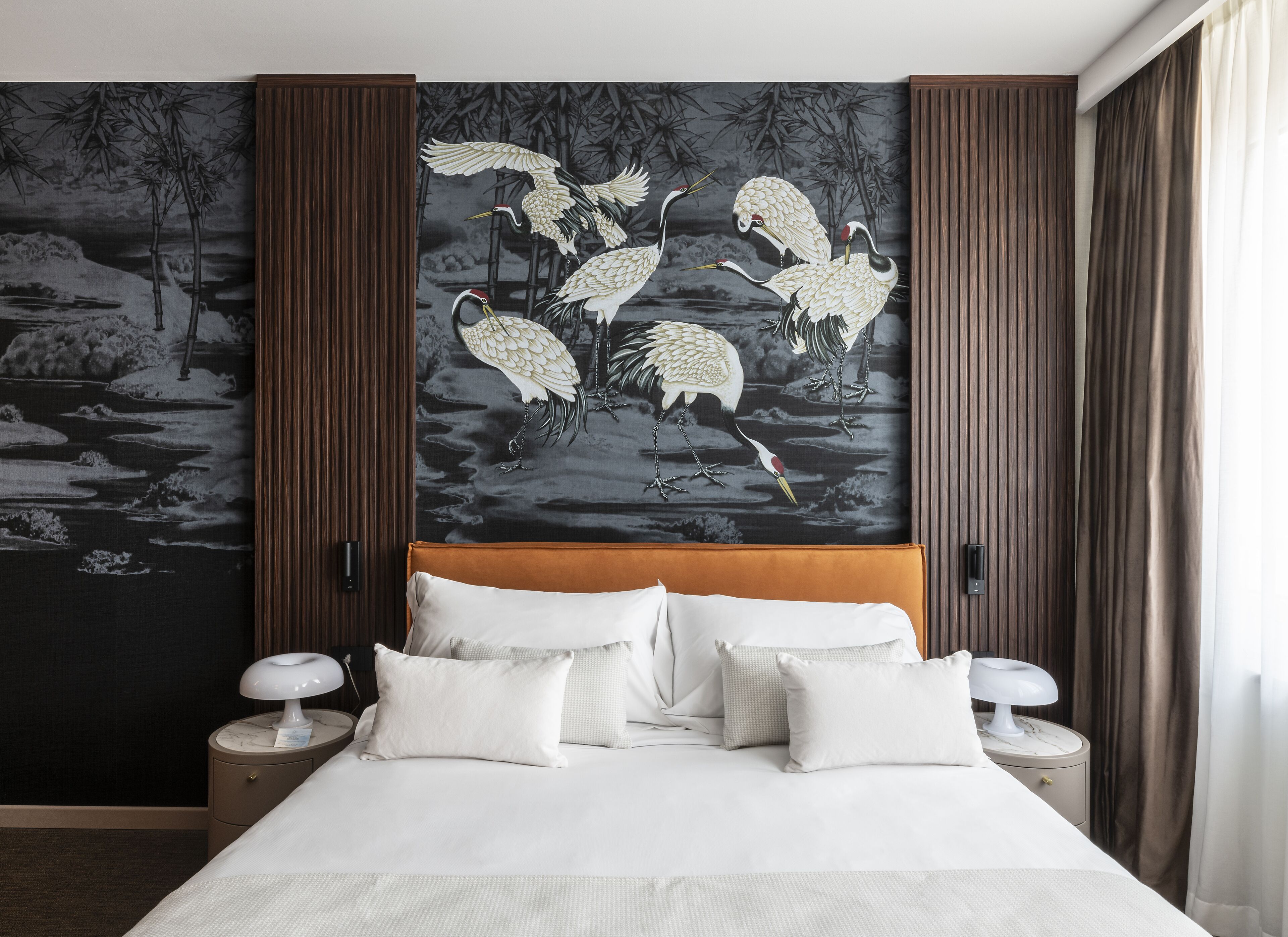 A hotel room with a large bed, two bedside tables, and a wall mural of cranes over water.