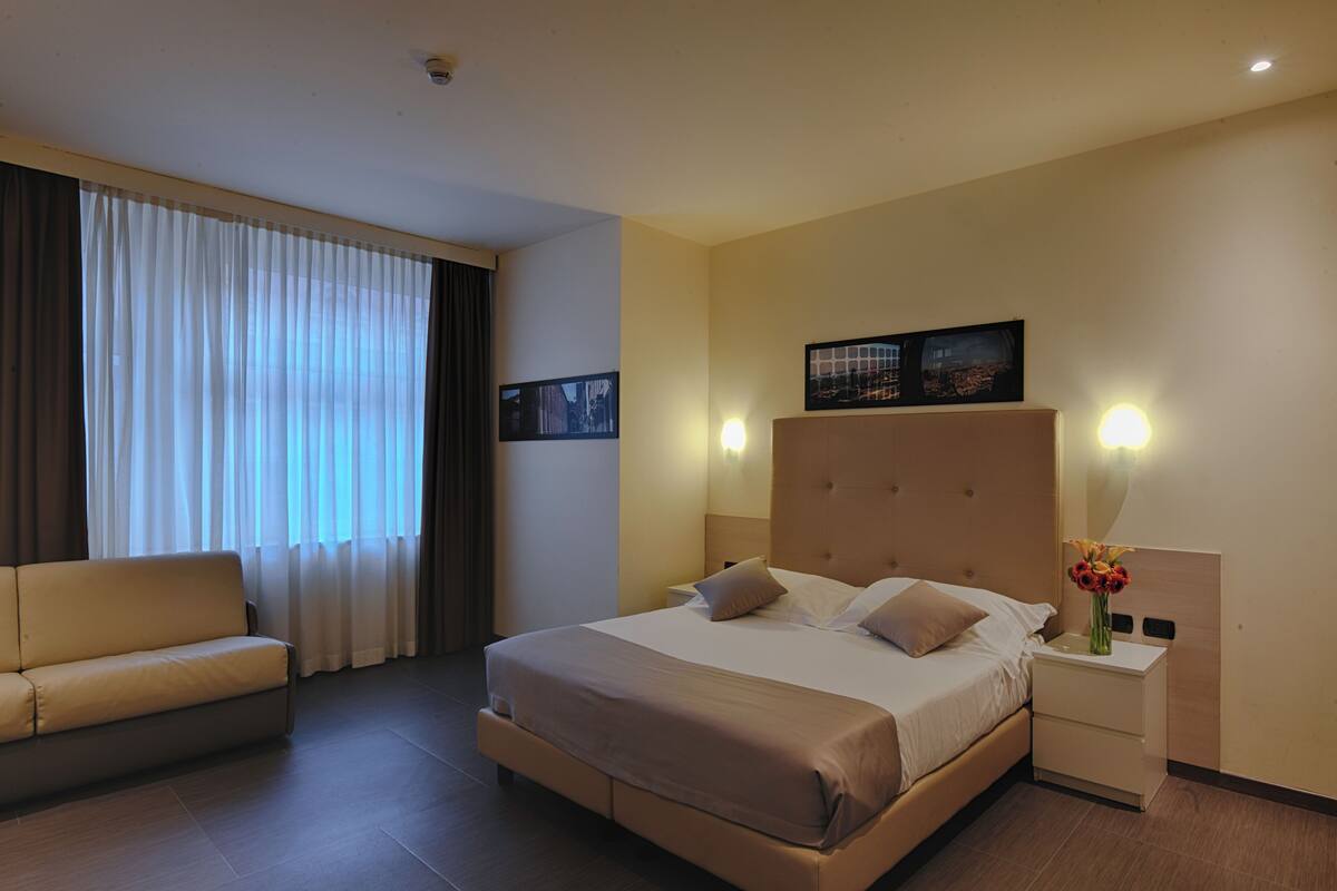 A hotel room with a large bed, a sofa, a nightstand with a vase of flowers, and two wall-mounted lamps.