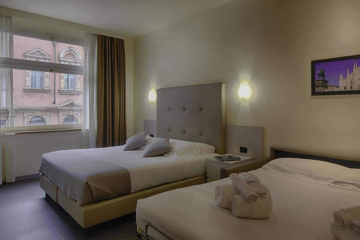 A hotel room with two beds, a large window with curtains, a wall-mounted light, and a framed picture.
