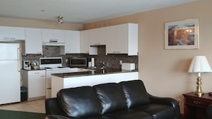 Een kleine keuken met witte kasten, een koelkast, een magnetron en een fornuis. Er staat een zwarte leren bank en een lamp op een houten bijzettafeltje.