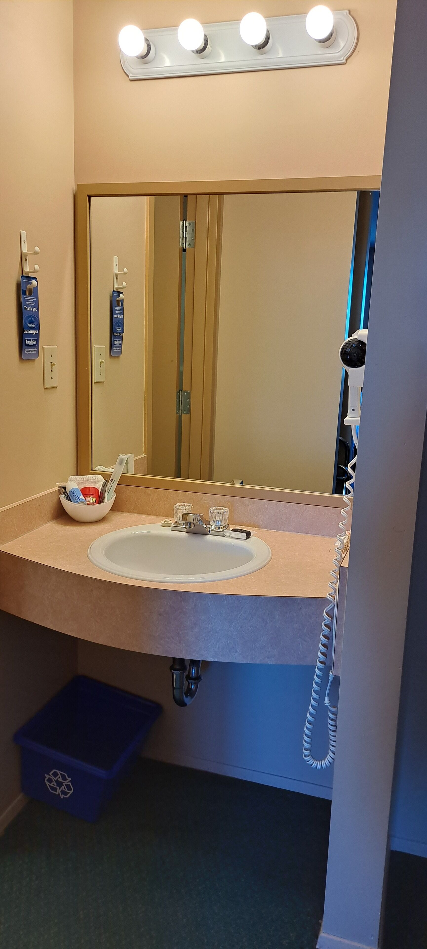 A bathroom with a sink, mirror, vanity lights, hand sanitizer dispensers, a hairdryer, and a trash bin.