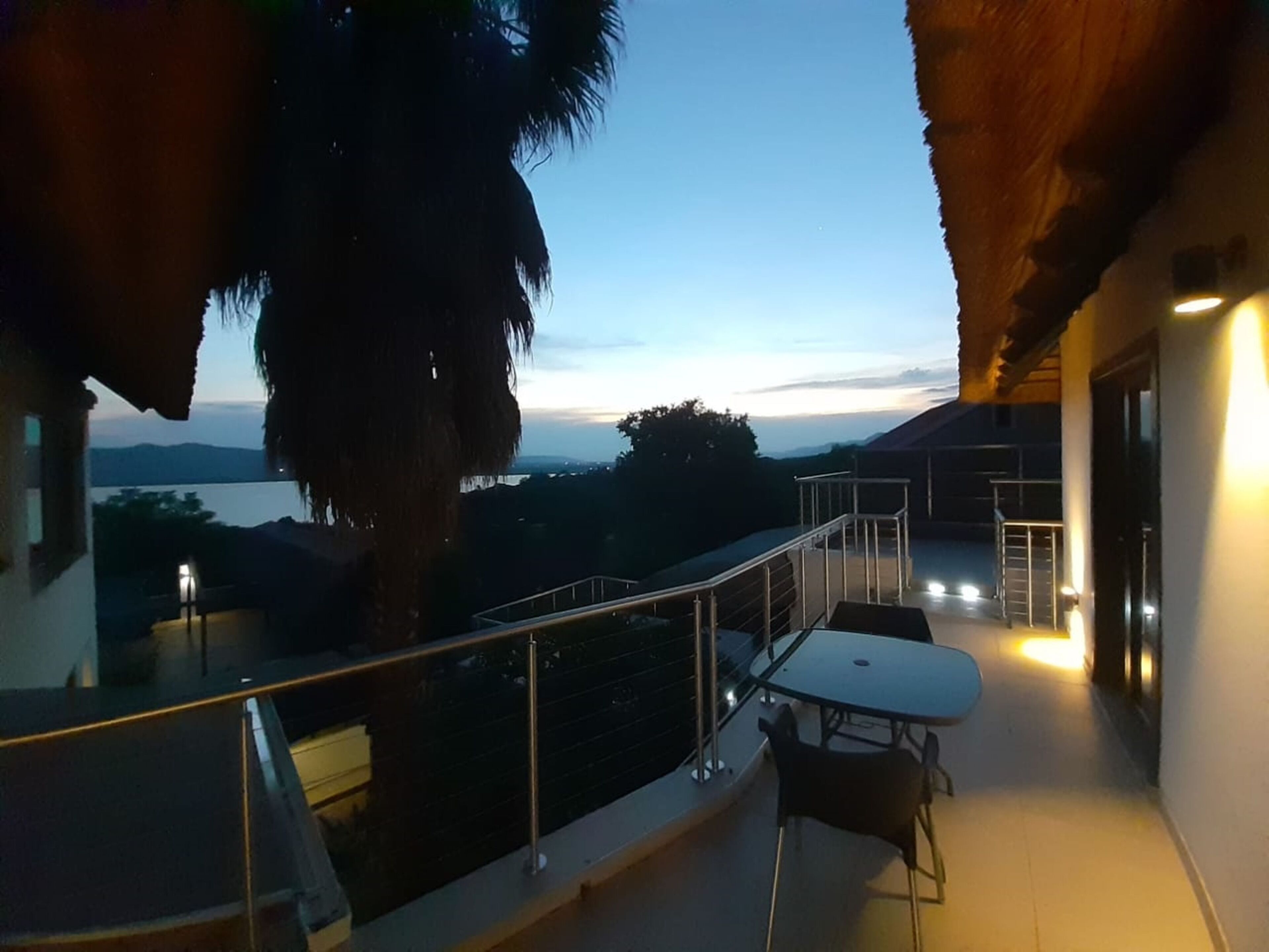 Un balcón con mesa y sillas, con vista a un paisaje pintoresco de un lago y montañas al anochecer.