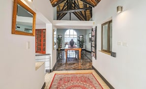 A hallway with a high ceiling, a red-framed mirror, a patterned rug, and a view through a large window. - Galagos Lodge (Hartbeespoort)