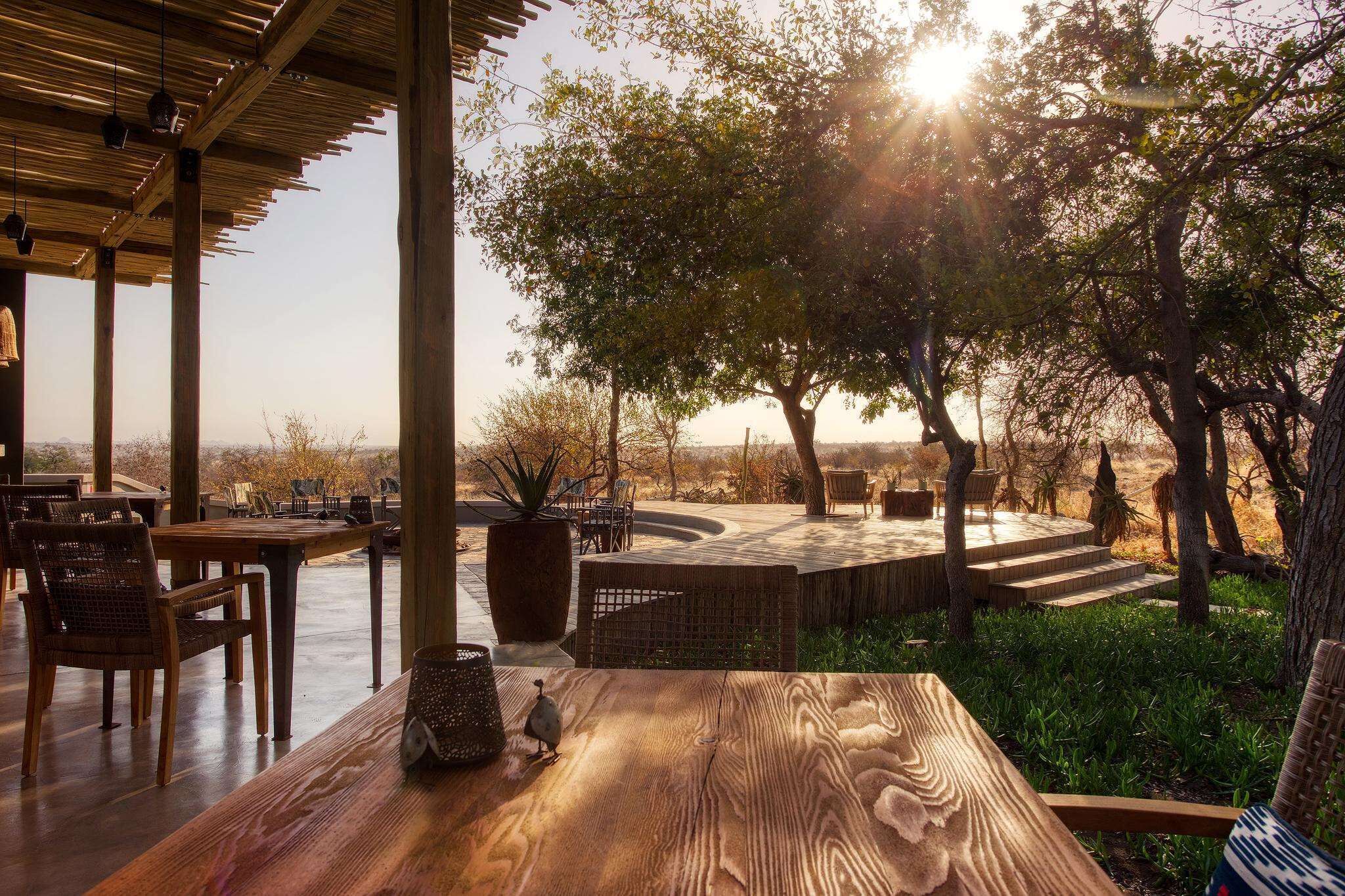 An outdoor dining area with wooden tables and chairs, surrounded by trees and a stone fire pit.