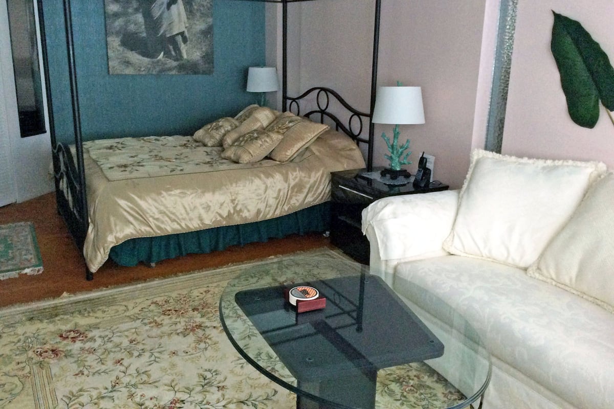 Un lit à baldaquin avec une couvre-lit à motifs floraux, un canapé blanc, une table basse en verre et une table de chevet avec une lampe et un téléphone.