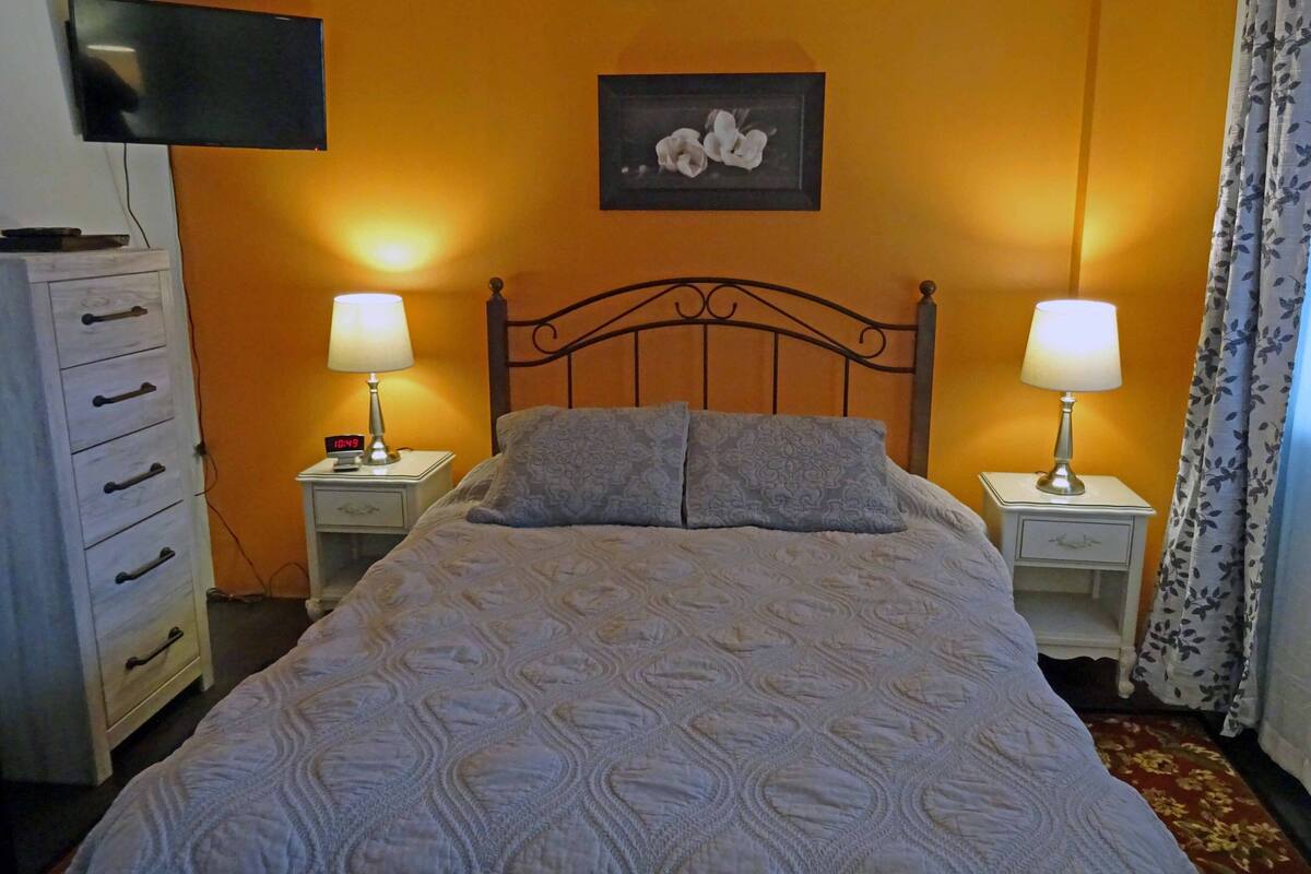 Une chambre à coucher avec un lit, deux tables de chevet avec des lampes, un téléviseur fixé au mur, une commode et un tableau encadré accroché au mur.
