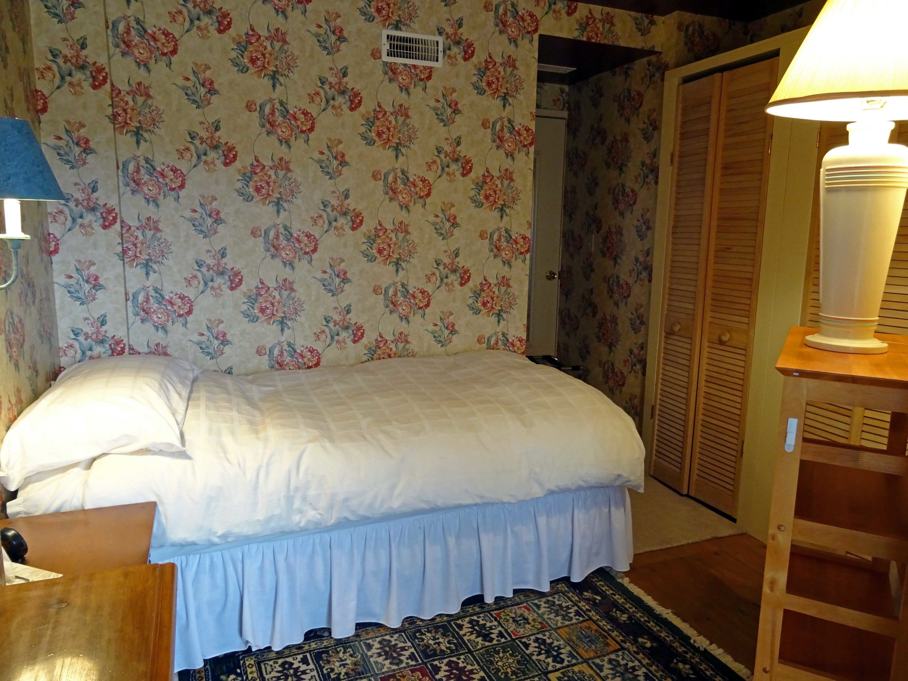 A bedroom with floral wallpaper, a bed with white bedding, a wooden nightstand, a lamp, and a patterned rug.