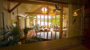 Il s'agit d'une pièce spacieuse avec un plafond haut, de grandes fenêtres et un lustre. On y trouve plusieurs tables et chaises disposées pour les repas ou les moments de convivialité.
