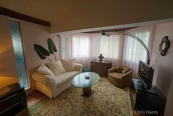 Una sala de estar con sofá, sillón, mesa de centro de vidrio y televisor sobre un mueble.