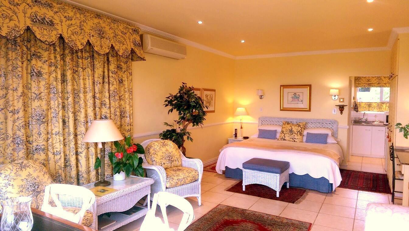 A hotel room with a bed, two chairs, a table, a lamp, a plant, and a small kitchen area.