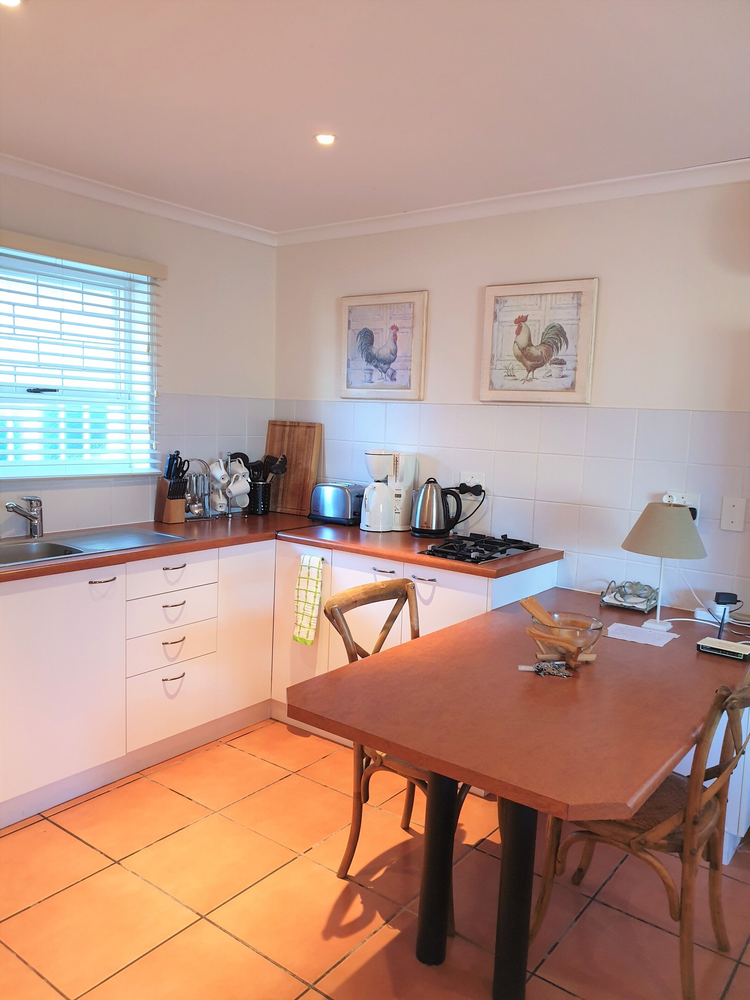 A kitchen with white cabinets, a wooden countertop, a sink, and a dining table with chairs. There are two framed pictures on the wall, a window with blinds, and a lamp on the countertop.