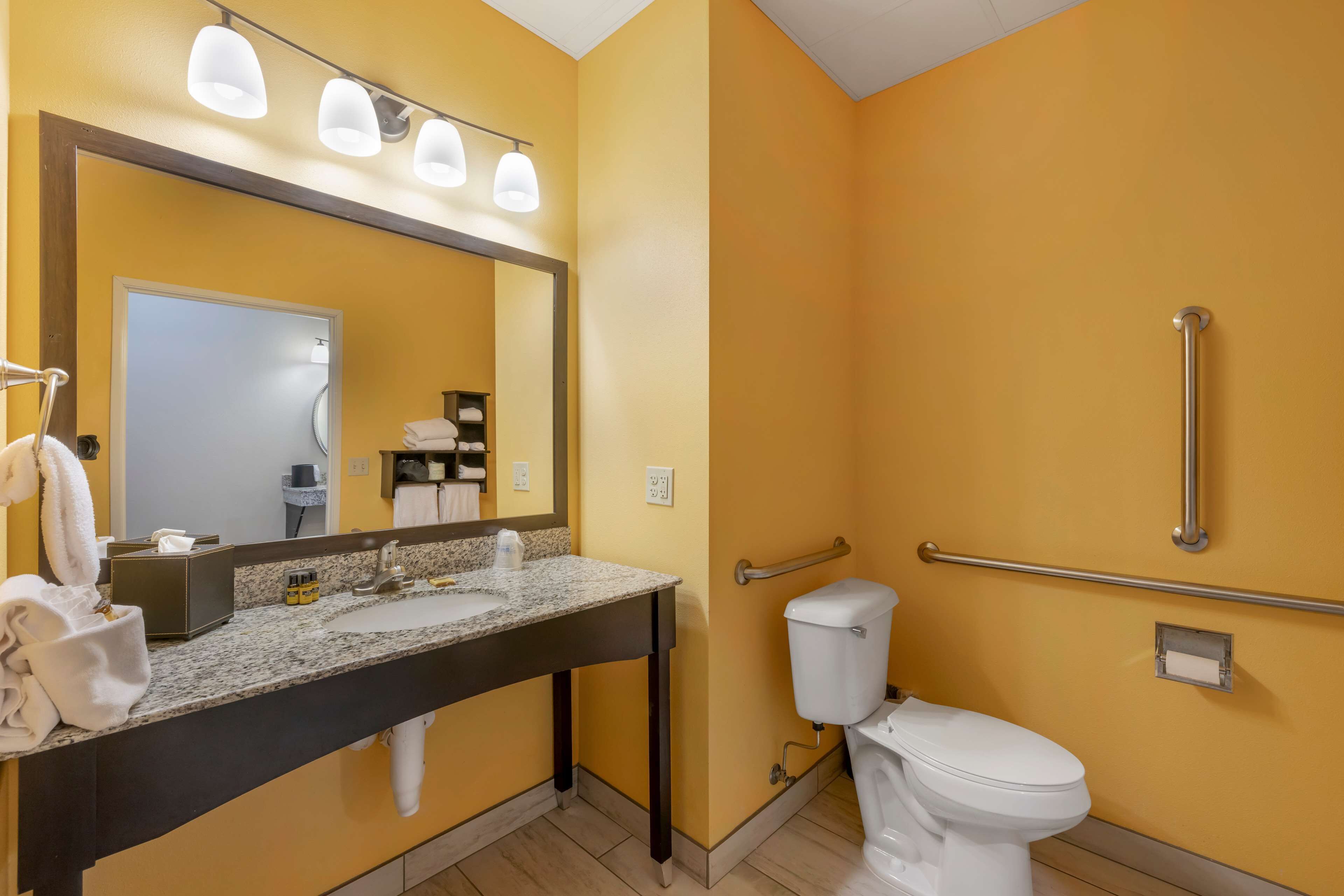 A bathroom with a yellow wall, a sink with a mirror, a toilet, and a handrail.