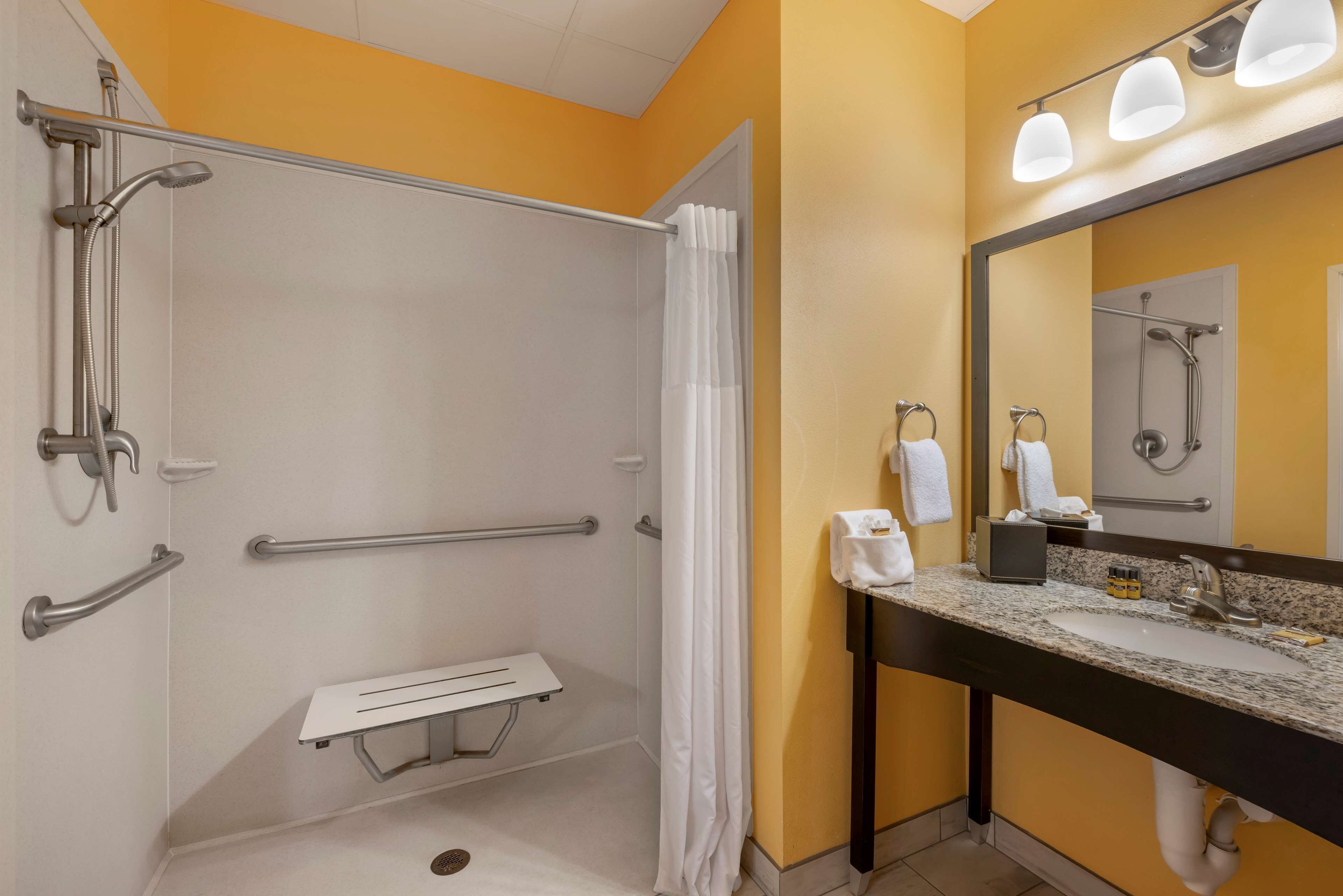 A bathroom with a shower, grab bars, a bench, a sink, and a mirror.