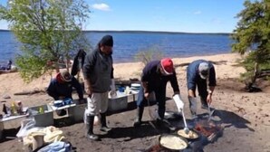 En gruppe mennesker laver mad udendørs ved en strand, med et vandområde i baggrunden.