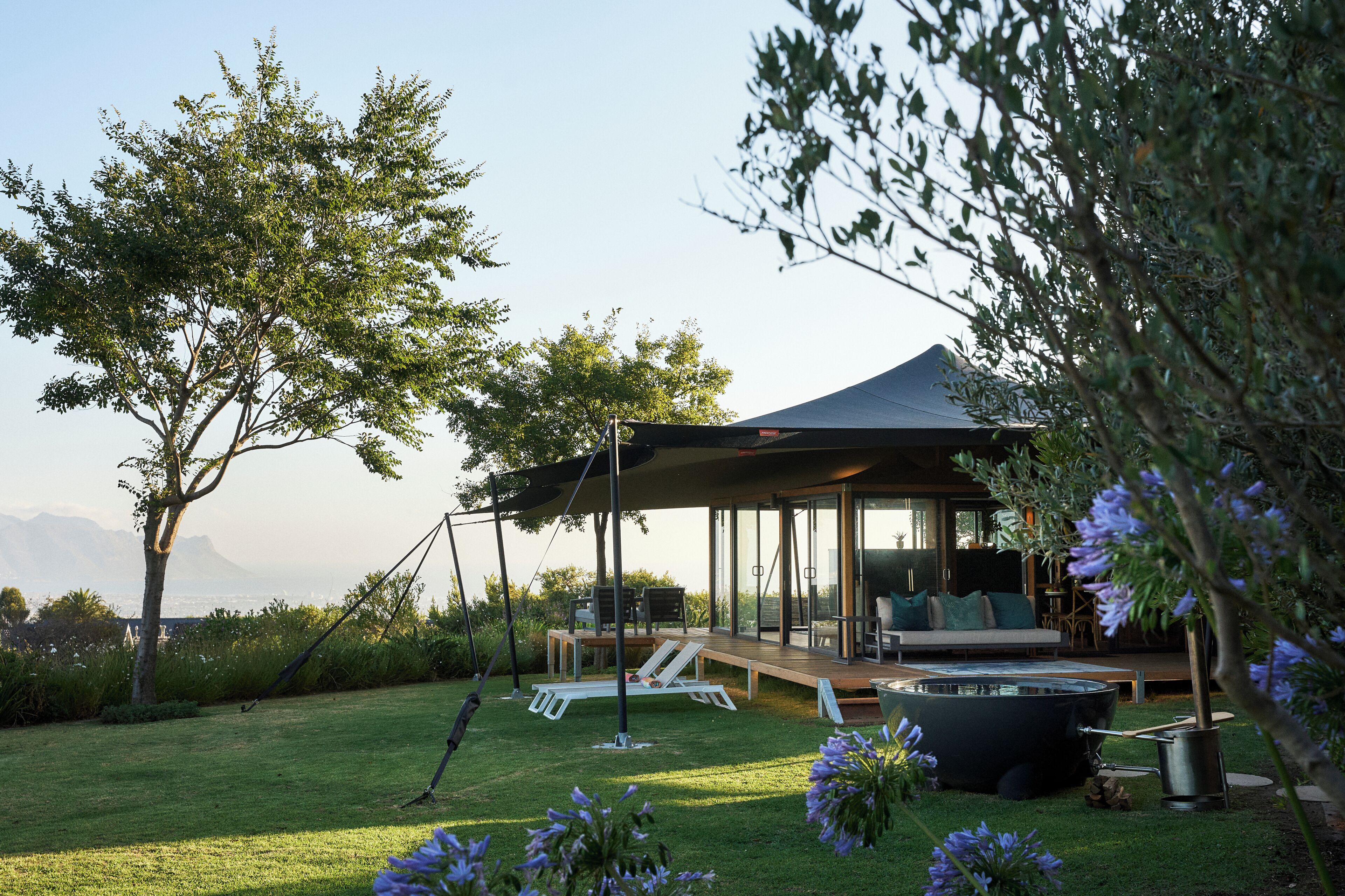 A modern outdoor lounge area with a glass enclosure, a hot tub, and a deck overlooking a grassy landscape with trees and mountains in the distance.