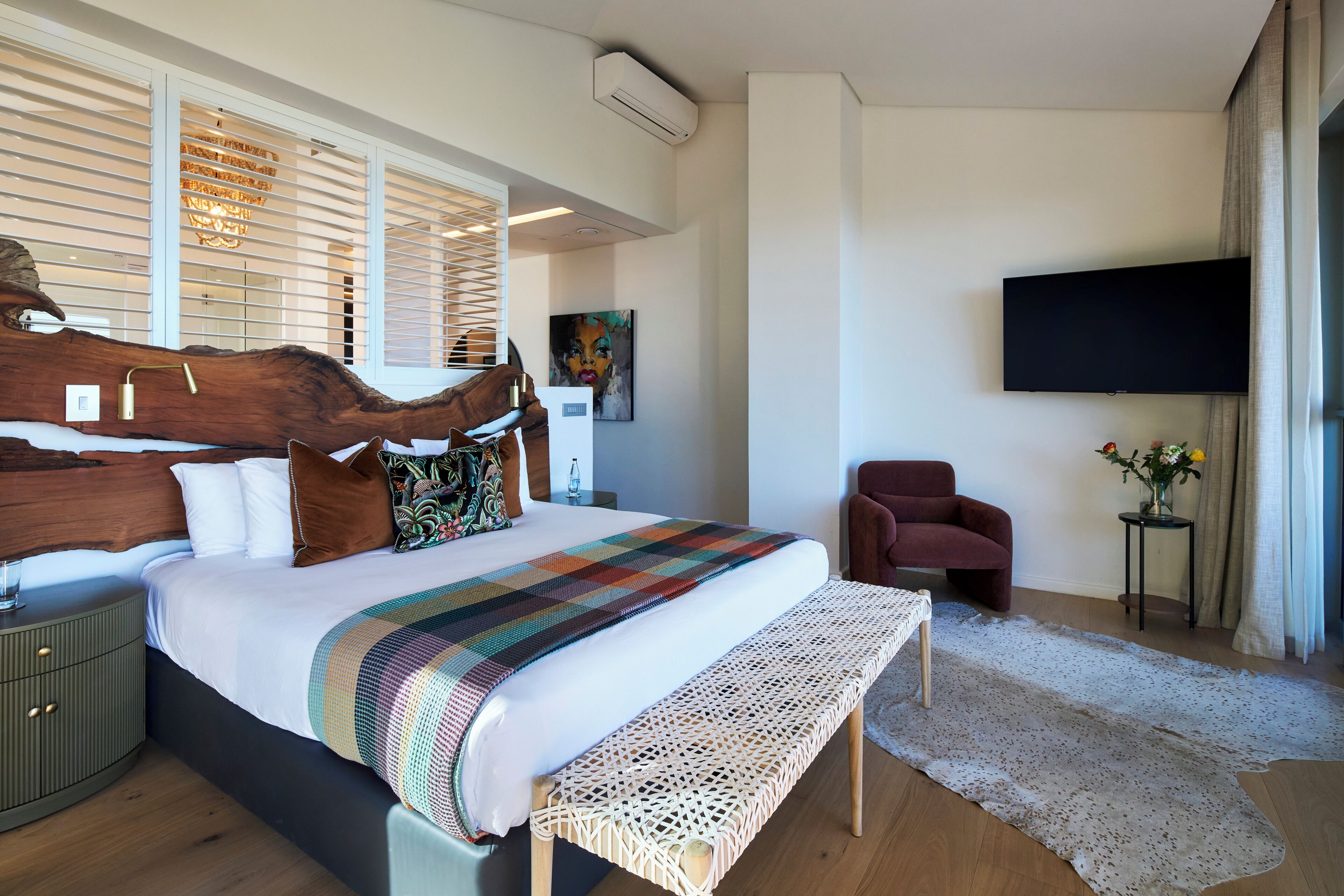 Moderni makuuhuone, jossa on suuri sänky, puinen pääty, taulutelevisio, punainen nojatuoli ja pieni sivupöytä, jossa on maljakko.