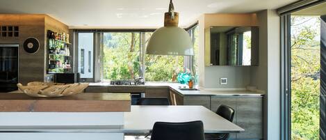 A modern kitchen with a large island, a dining area with a table and chairs, and a view of greenery through large windows.