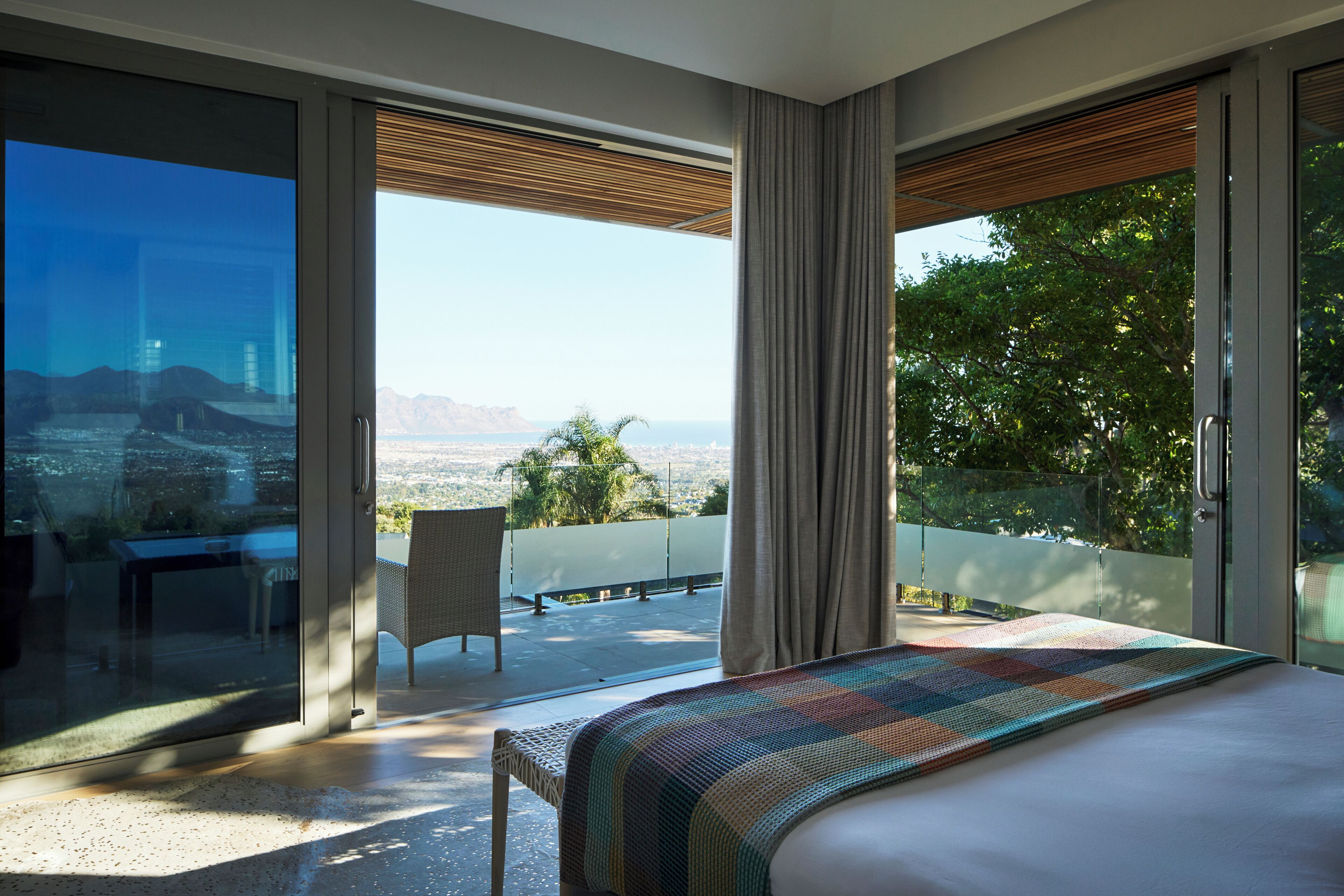 A modern hotel room with a large bed, a view of the cityscape, and a balcony with a table and chair.