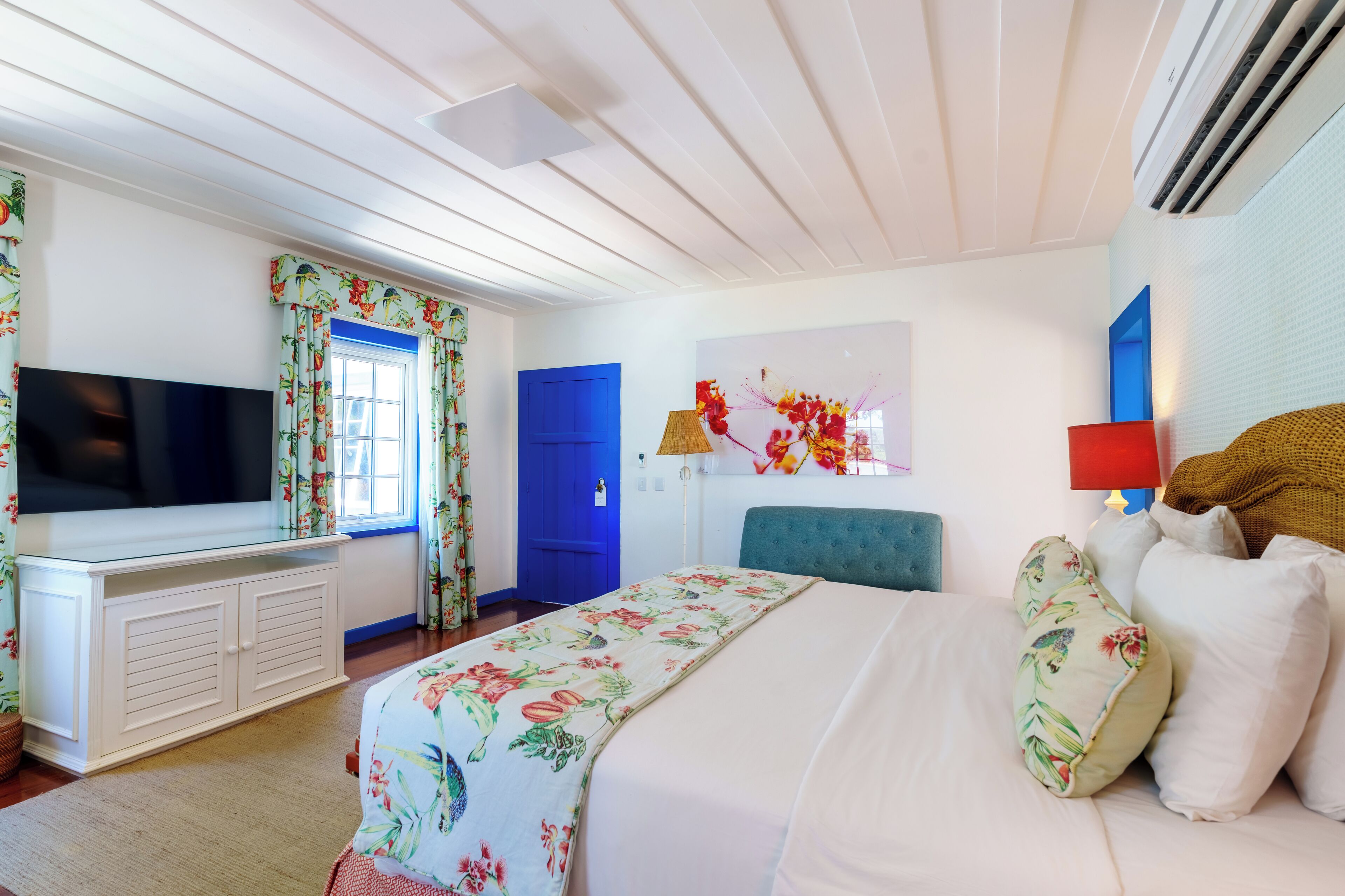 A bedroom with a bed, a TV, a blue door, and a window with floral curtains.