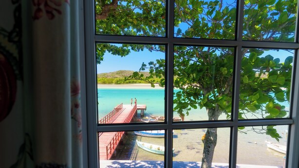 A view through a window with a grid pattern, displaying a beach with boats, a wooden pier, and a tree. - Hotel Solar do Arco (Cabo Frio)