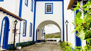A white building with blue trim and a tiled roof, a stone pathway leading to an archway, and red flowers in the foreground. - Hotel Solar do Arco (Cabo Frio)