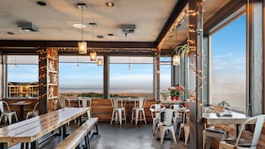 Un restaurante con mesas y sillas de madera, amplios ventanales con vistas y luces colgantes.